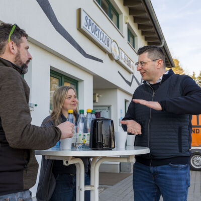 Bild vergr&ouml;&szlig;ern: Der Landrat spricht vor dem Geb&auml;ude des Sportclubs Syrau mit zwei Vereinsverantwortlichen an einem Stehtisch. Auf dem Tisch stehen Tassen, eine Kanne und Wasserflaschen. Im Hintergrund ist ein Anh&auml;nger zu sehen.