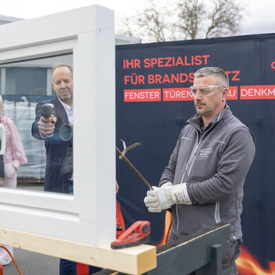 Bild vergr&ouml;&szlig;ern: Ein Mitarbeiter mit Schutzbrille und Handschuhen steht neben einem eingebauten Fenster f&uuml;r eine Brandschutz-Demonstration im Freien.