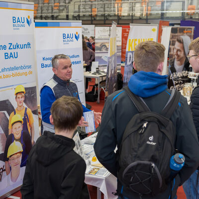 Bild vergr&ouml;&szlig;ern: Mehrere Jugendliche mit Rucks&auml;cken stehen an einem Messestand und sprechen mit einem Aussteller. Hinter ihm stehen gro&szlig;e Werbebanner von BAU Bildung Sachsen mit Informationen zu Ausbildung und Studium im Bauwesen. Die Szene zeigt ein Beratungsgespr&auml;ch in offener Messeatmosph&auml;re.