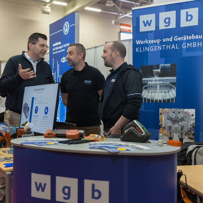 Bild vergr&ouml;&szlig;ern: Drei M&auml;nner, darunter der Landrat stehen an einem Messestand mit blauen Werbetafeln und unterhalten sich. Auf dem Stand liegen Informationsmaterialien und technische Anschauungsobjekte. Im Hintergrund sind weitere Messest&auml;nde und Besucherinnen und Besucher zu erkennen.