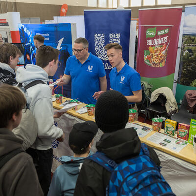 Bild vergr&ouml;&szlig;ern: Eine Gruppe junger Besucher steht vor einem Messestand, an dem zwei Aussteller in blauen Poloshirts informieren. Auf dem Tisch liegen Flyer und verschiedene Produktverpackungen. Im Hintergrund sind weitere Banner und St&auml;nde in der Halle zu sehen.