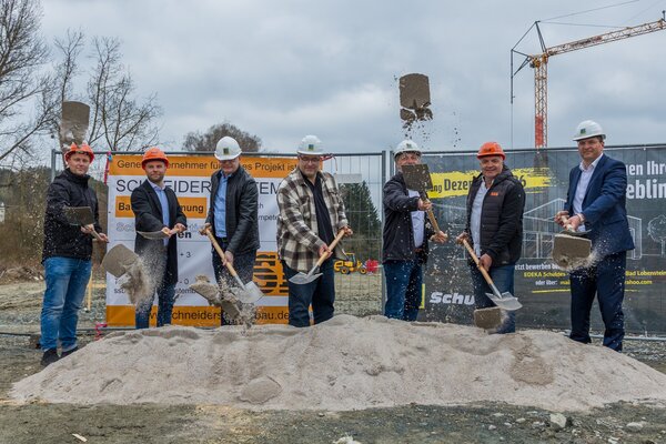 Bild vergr&ouml;&szlig;ern: Sieben M&auml;nner stehen mit Helmen und Spaten vor einem Sandhaufen auf der Baustelle. Im gleichen Moment werfen sie Sand f&uuml;r den symbolischen Spatenstich in die Luft. Hinter ihnen h&auml;ngen Bauzaunbanner, im Hintergrund ragt ein Kran in den bew&ouml;lkten Himmel.