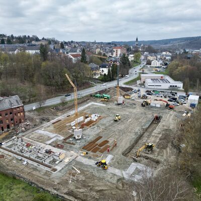 Bild vergr&ouml;&szlig;ern: Drohnenaufnahme einer gro&szlig;en Baustelle am Rand von Adorf. Auf dem weitl&auml;ufigen Gel&auml;nde sind Erdarbeiten, Fundamente, Baumaschinen und zwei Kr&auml;ne zu sehen. Im Hintergrund liegen Stra&szlig;e, Wohnh&auml;user und die Silhouette der Stadt unter einem grauen Himmel.