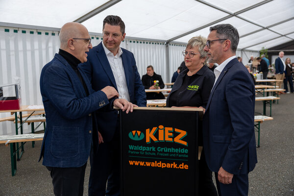 Bild vergr&ouml;&szlig;ern: Vier Personen stehen w&auml;hrend des Festaktes zum 35-j&auml;hrigen Bestehen des KiEZ Waldpark Gr&uuml;nheide in einem Festzelt zusammen und unterhalten sich an einem Rednerpult mit KiEZ-Logo. Zu sehen sind Joachim Otto, Landrat Thomas Hennig, Sylvia Ott und KiEZ-Pr&auml;sident S&ouml;ren Voigt.