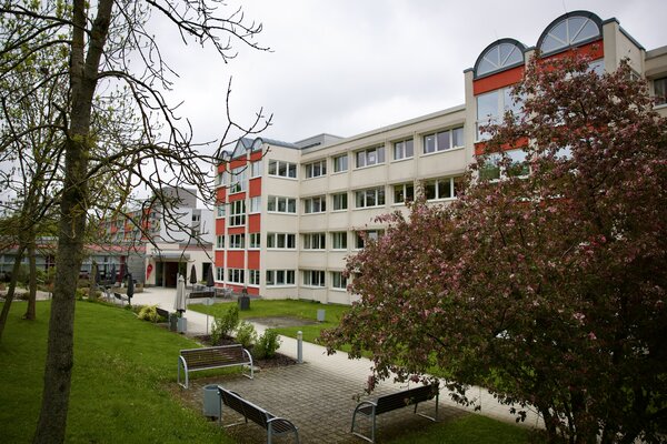 Bild vergr&ouml;&szlig;ern: Au&szlig;enansicht der AWO Tagespflege Panoramablick in Auerbach mit hellem Geb&auml;ude, Gr&uuml;nfl&auml;chen, Sitzb&auml;nken und bl&uuml;hendem Baum im Vordergrund.