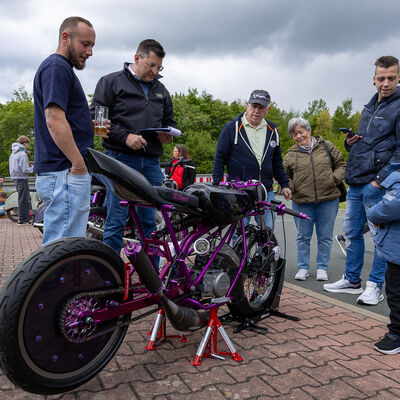 Bild vergr&ouml;&szlig;ern: Mehrere M&auml;nner und Frauen sowie Landrat Thomas Hennig stehen im Halbkreis um ein violettes, tiefergelegtes Motorrad mit auff&auml;llig breitem Hinterreifen. Ein Besucher mit Bierglas in der Hand betrachtet die Maschine, andere unterhalten sich.
