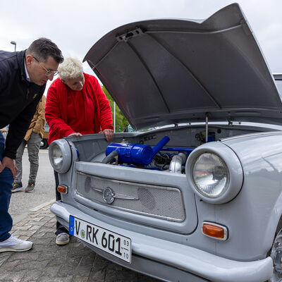 Bild vergr&ouml;&szlig;ern: Landrat Thomas Hennig und Anja Strobel-Schaller beugen sich beim Rundgang &uuml;ber den ge&ouml;ffneten Motorraum eines wei&szlig;en Trabant. Im Motorraum ist ein auff&auml;llig blauer Ansaugschlauch zu sehen.