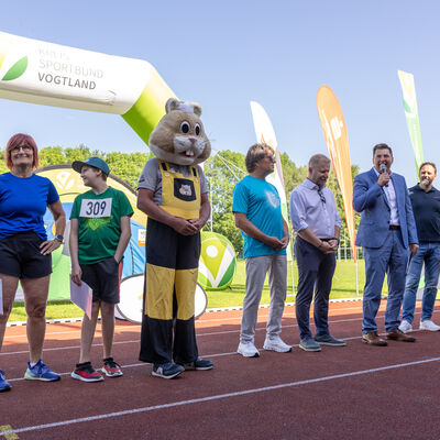 Bild vergr&ouml;&szlig;ern: Eine Reihe von Personen steht auf der Laufbahn unter einem gr&uuml;n-wei&szlig;en aufblasbaren Bogen mit der Aufschrift &raquo;Kreissportbund Vogtland&laquo;. Von links: eine Frau im blauen Sportshirt, ein Junge im gr&uuml;nen Trikot mit Startnummer 309, ein Maskottchen im Hamster-Kost&uuml;m mit gelber Latzhose, mehrere M&auml;nner in Freizeit- und Anzugkleidung. Ein Mann im hellblauen Anzug spricht in ein Mikrofon. Sonniger Tag, im Hintergrund Werbefahnen und B&auml;ume.