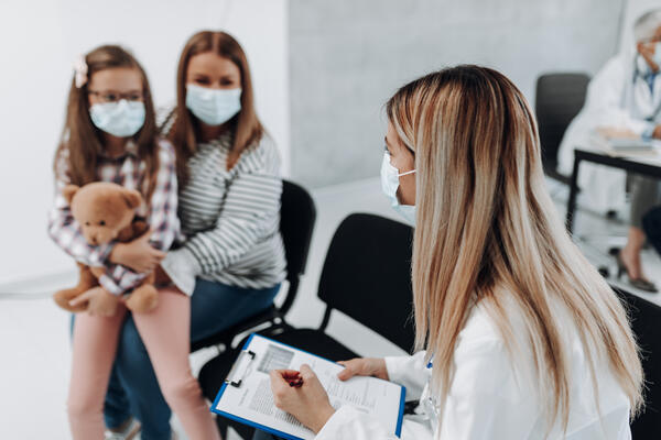 Bild vergr&ouml;&szlig;ern: Das Bild zeigt ein Kind auf dem Scho� seiner Mutter. Beide sitzen einer �rztin gegen�ber. Diese h�lt ein Formular in der Hand und arbeitet dieses nach Stichpunkten ab.