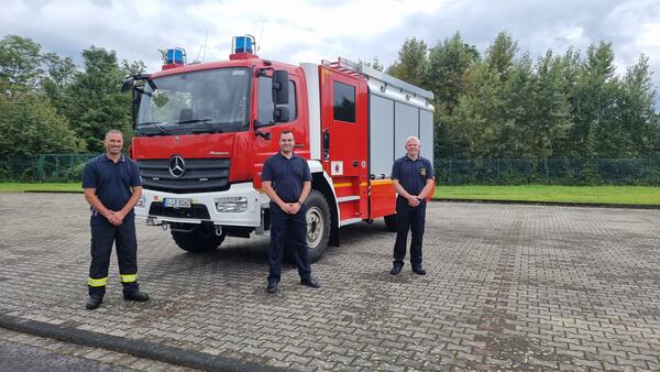 Bild vergr&ouml;&szlig;ern: Das Bild zeigt drei M&auml;nner in blauer Uniform vor einem roten Feuerwehrauto.
