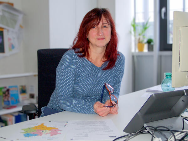 Bild vergr&ouml;&szlig;ern: Das Bild zeigt die Koordinatorin f&uuml;r Schulsozialarbeit im Landratsamt vogtlandkreis Birgit Stief. Sie tr&auml;gt einen hellblauen Pullover und sitzt an ihrem Schreibtisch. Sie l&auml;chelt in die Kamera und h&auml;lt dabei ihre Brille in der Hand.