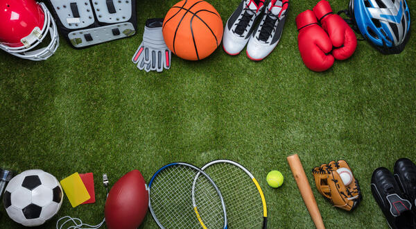 Bild vergr&ouml;&szlig;ern: Das Bild zeigt eine Auswahl verschiedener Sportgegenst�nde auf einem gr�nen Rasen. Darunter sind ein Basketball, Federballschl�ger und Fu�ballschuhe.
